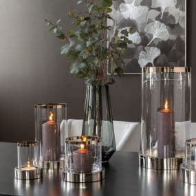 Kerzen in Glasbehältern auf einem Tisch, dahinter Vase mit grünen Zweigen und Schwarz-Weiß-Gemälde.