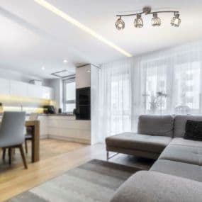 Modernes Wohnzimmer mit grauem Sofa, Essbereich und offener Küche. Helle Vorhänge und Deckenspots.