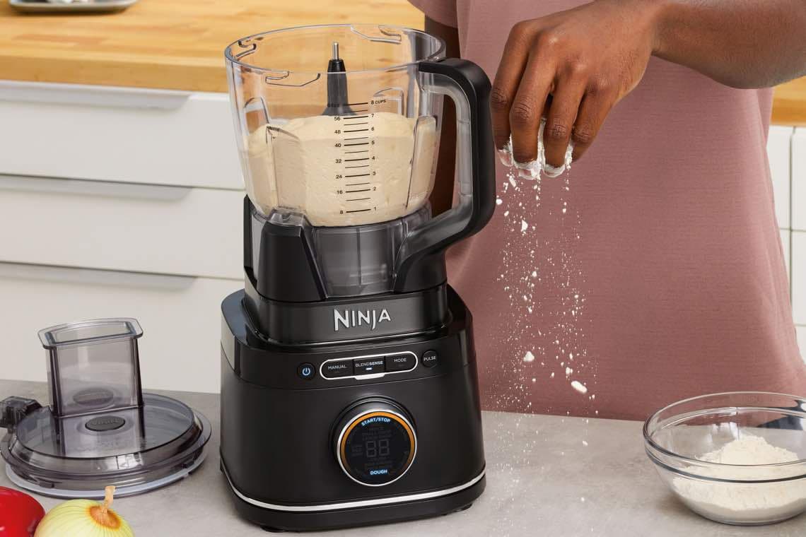 Hand streut Mehl in Küchenmaschine von Ninja, daneben Schüssel mit Mehl und Zutaten auf der Arbeitsplatte.