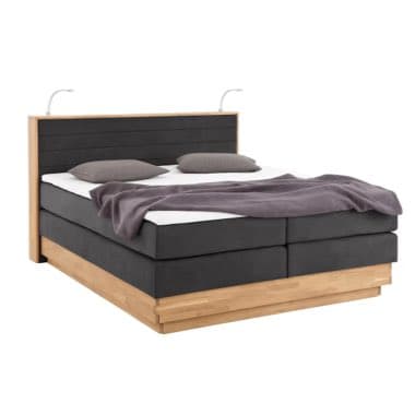 Doppelbett mit hohem Kopfteil, grauer Decke und Kissen, auf hellem Sockel, mit zwei flexiblen Leselampen am Kopfende