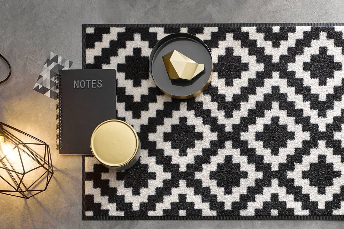 Notizbuch mit Aufdruck NOTES auf Teppich mit schwarz-weißem Rautenmuster, daneben Deko und Lampe