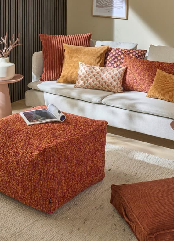 Beiger Sofa mit orangefarbenen Kissen, passender Pouf mit aufgeklapptem Magazin in einem hellen Wohnzimmer