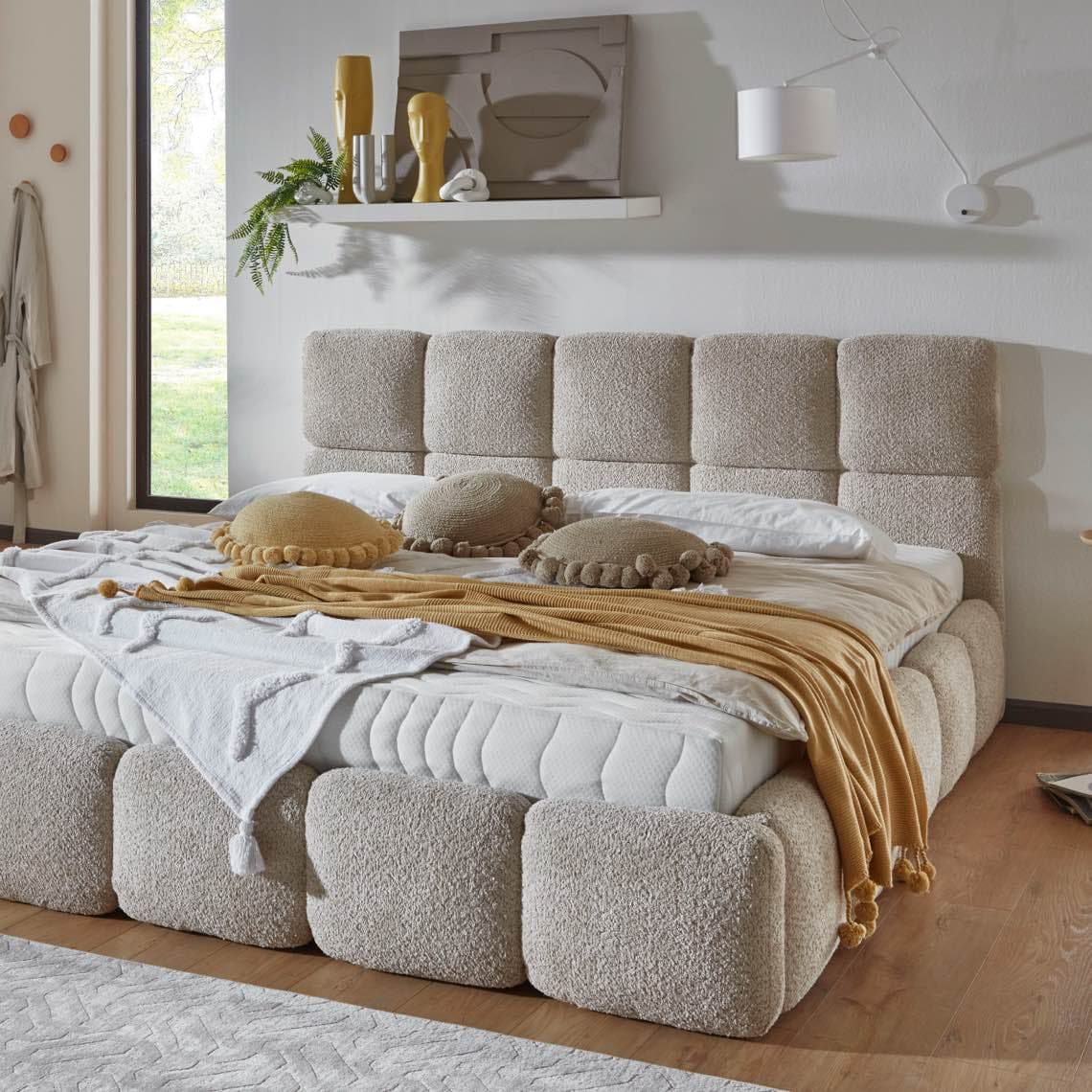 Großes Bett mit gepolstertem Kopfteil, runden Zierkissen und gelber Decke in hellem Schlafzimmer mit Fenster und Wandregal