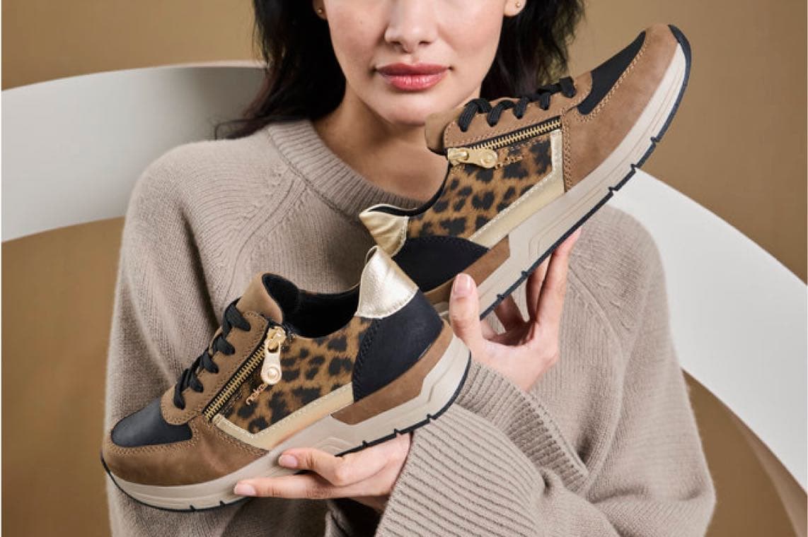 Frau im beigen Pullover hält zwei braune Sneaker mit Leopardenmuster und goldenen Details vor der Brust
