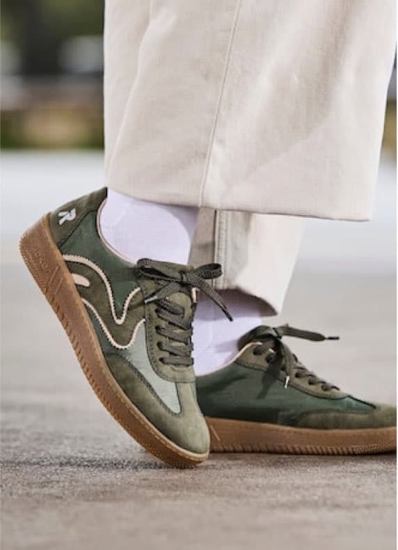 Person trägt olivgrüne Sneakers mit Wellenmuster und weißen Socken zu hellen Hosen, ein Fuß leicht angehoben