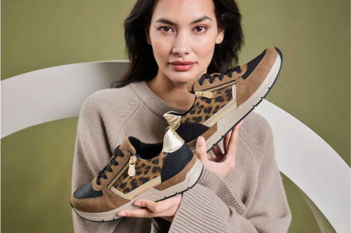 Frau im beigen Pullover hält Paar braun-schwarze Sneaker mit Leopardenmuster vor grünem Hintergrund