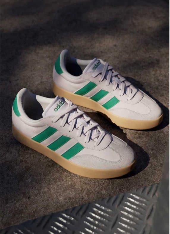 Weiße Adidas Sneaker mit grünen Streifen und beiger Sohle liegen schräg nebeneinander auf grauem Boden