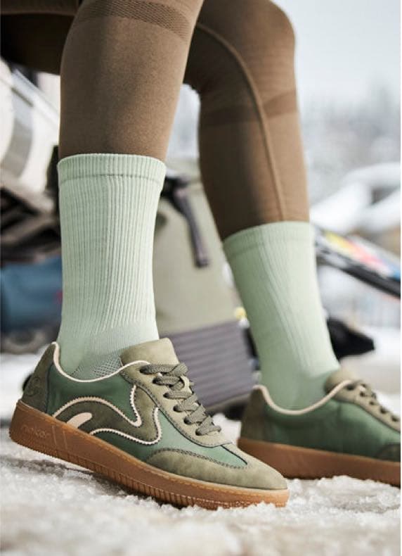 Person mit hellgrünen Socken und grünen Sneakern steht in brauner Leggings auf Schnee, Winterausrüstung im Hintergrund