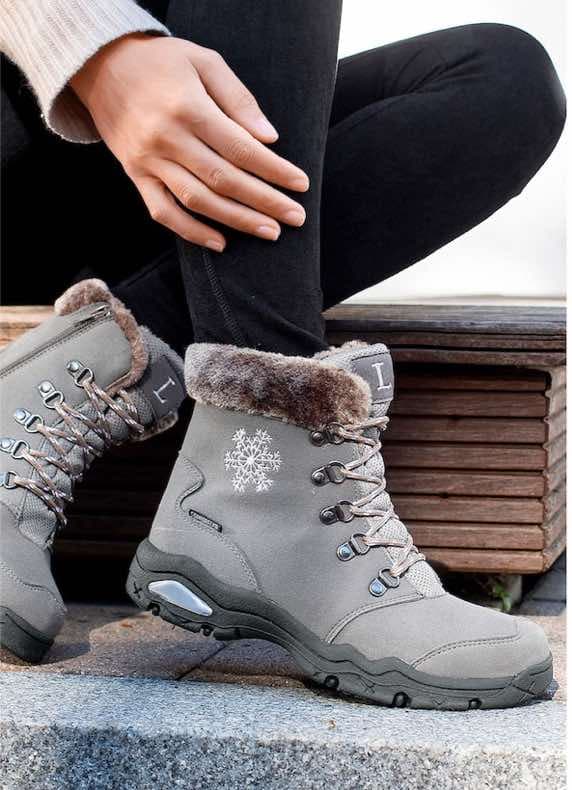 Graue Winterstiefel mit Schneeflockenmotiv, seitlichem Reißverschluss und Fellrand an den Füßen einer sitzenden Person