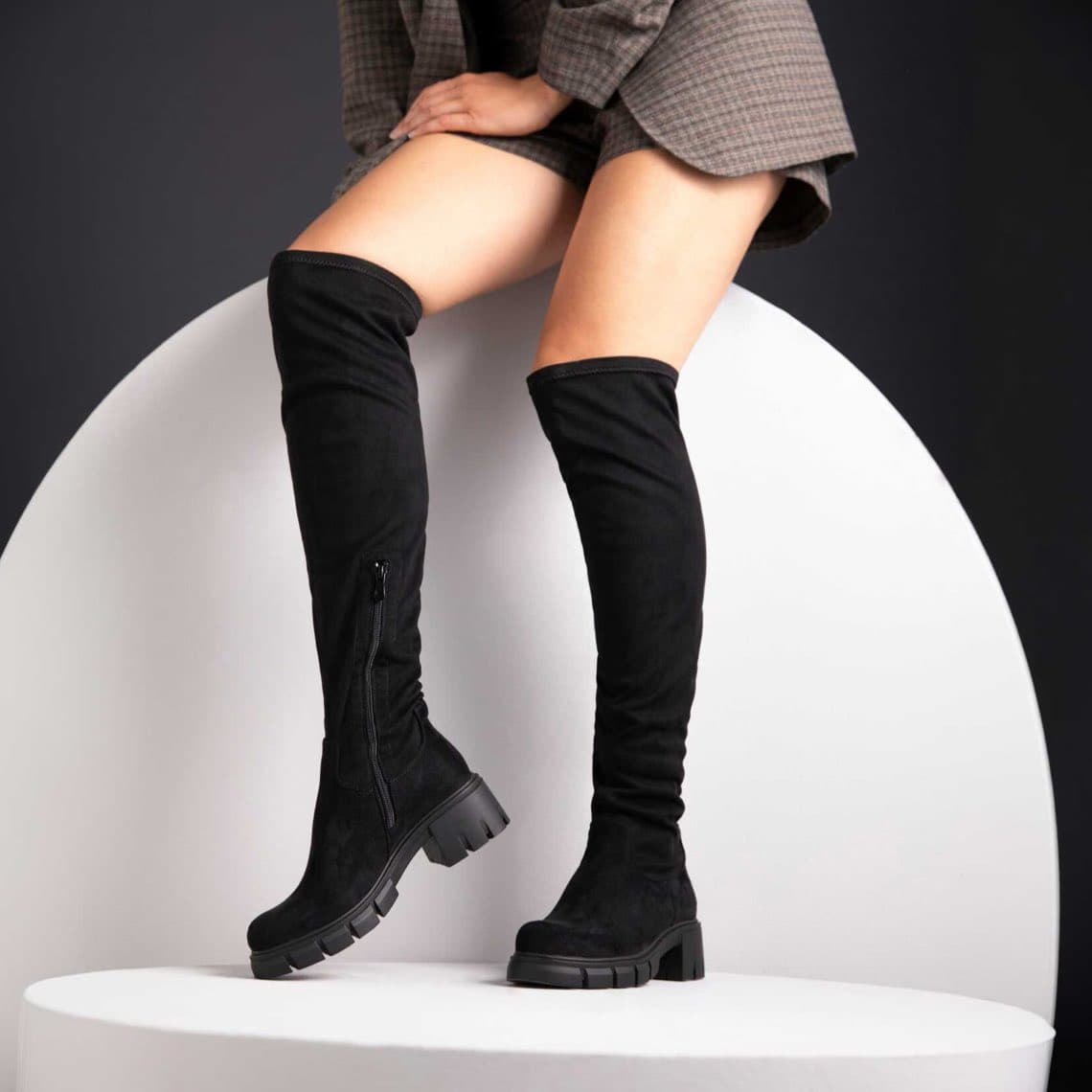 Frau sitzt auf weißem Podest und trägt schwarze Overknee-Stiefel mit dicker Profilsohle