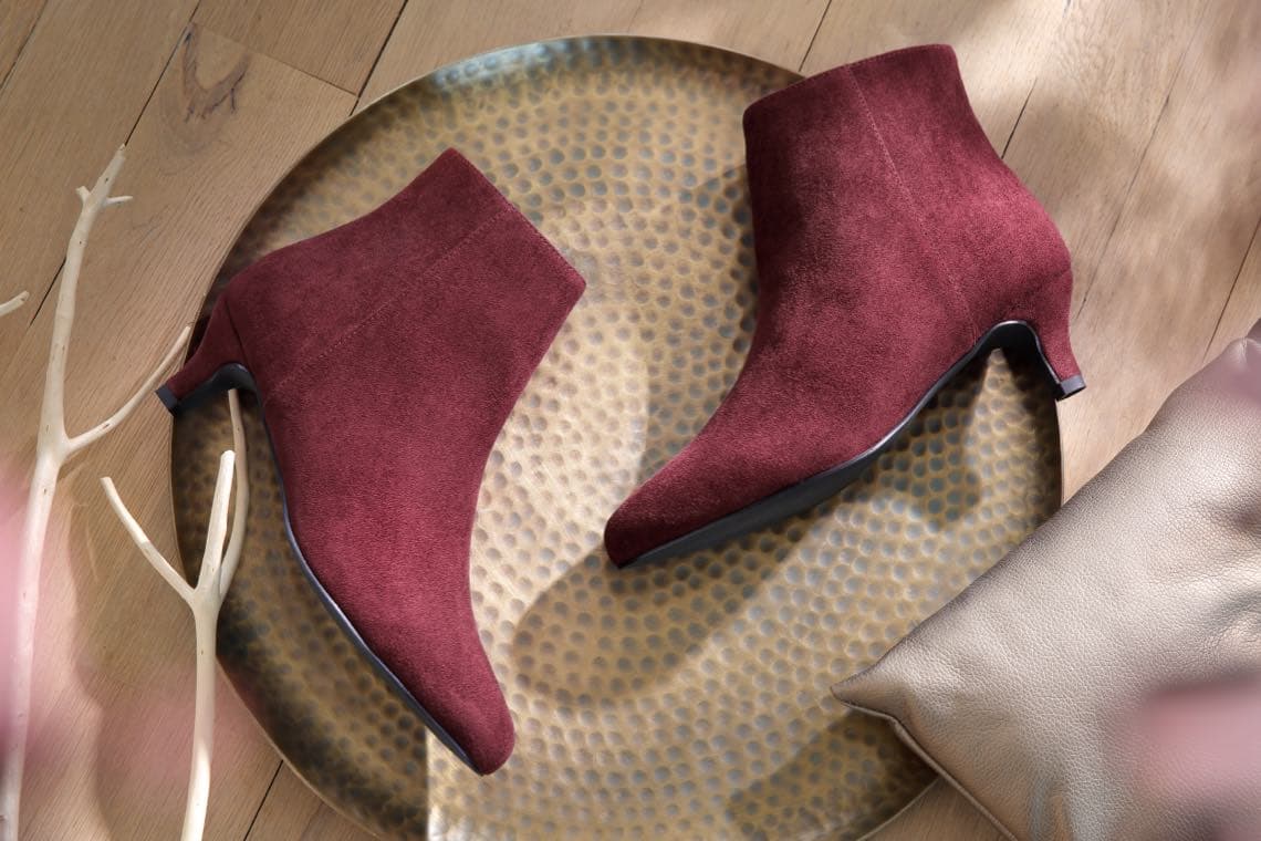 Bordeauxrote Stiefeletten mit kleinem Absatz auf runder Metallplatte, daneben Dekoast und Kissen auf Holzfußboden