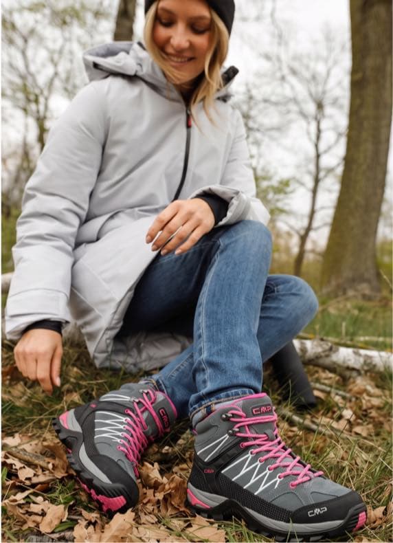 Frau im Wald sitzt auf einem Baumstamm und zeigt ihre knöchelhohen Wanderschuhe mit pinken Schnürsenkeln