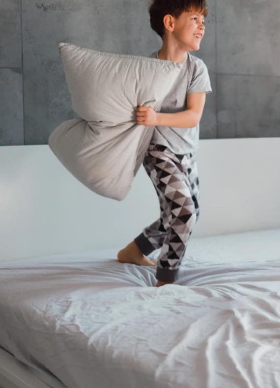 Junge im Schlafanzug springt auf dem Bett und hält ein großes Kopfkissen in den Armen