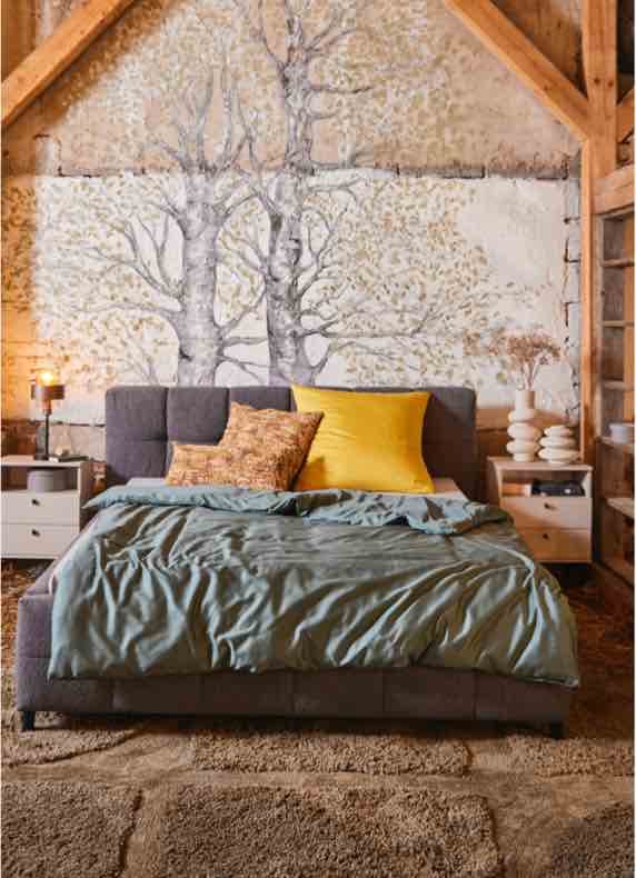 Doppelbett mit grüner Decke und gelben Kissen vor Wandbild mit Bäumen, flankiert von zwei Nachttischen