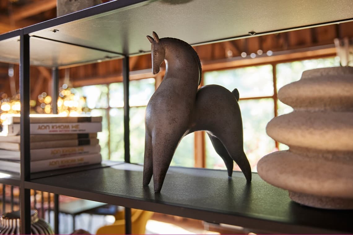 Dekorative Pferdefigur auf Regalboden, dahinter Bücherstapel und unscharfer Wohnraum mit großen Fenstern