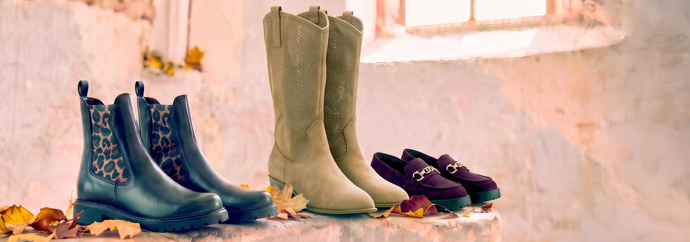 Fünf Paar Damenstiefel in Blau, Beige, Lila und Grün stehen auf einer Fläche zwischen herbstlichen Blättern.