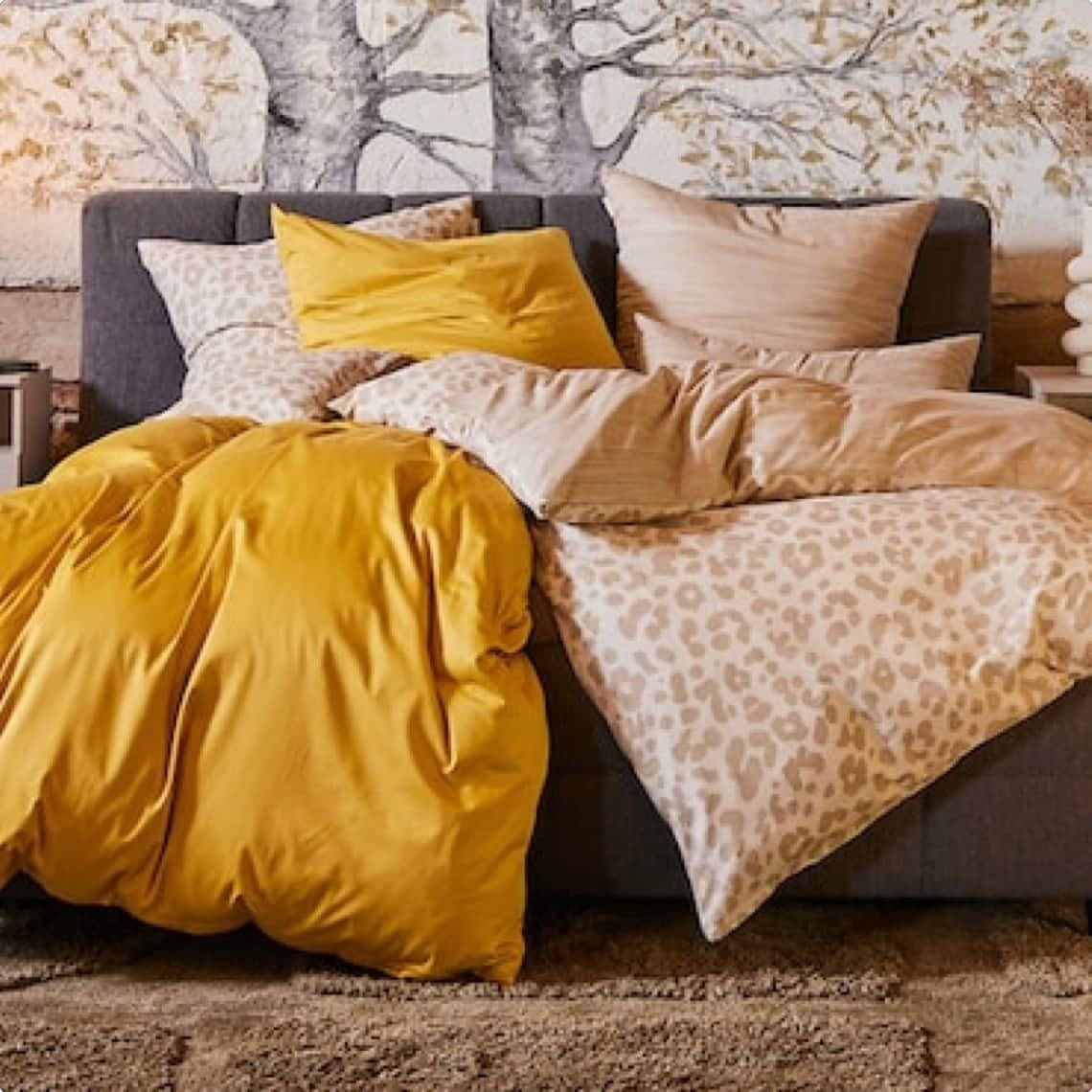 Doppelbett mit gelber und gemusterter Bettdecke, mehreren Kissen und Baum-Motiv an der Wand im Hintergrund
