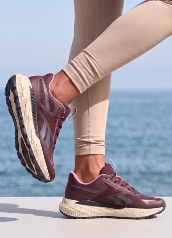 Person in beige Leggings trägt bordeauxfarbene Laufschuhe und steht vor dem Meer
