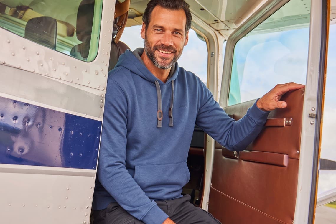 Mann im blauen Hoodie sitzt lächelnd in der offenen Tür eines kleinen Flugzeugs