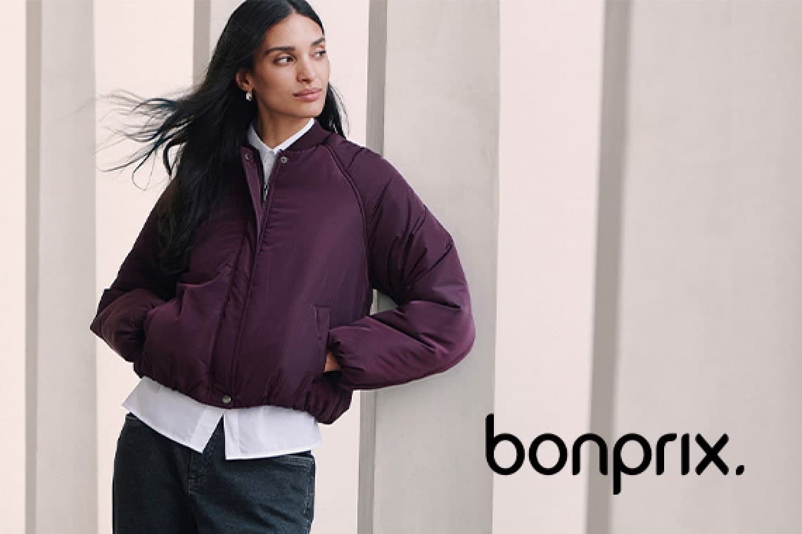 Frau in dunkler Steppjacke lehnt an heller Säule, Hände in den Taschen, bonprix Logo unten rechts