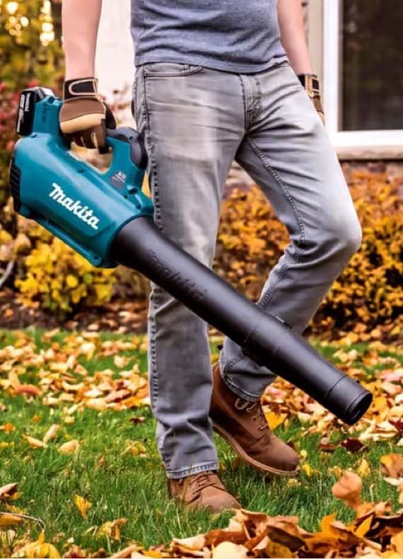 Person in Jeans hält Laubbläser von Makita, steht im herbstlichen Garten mit buntem Laub auf dem Rasen