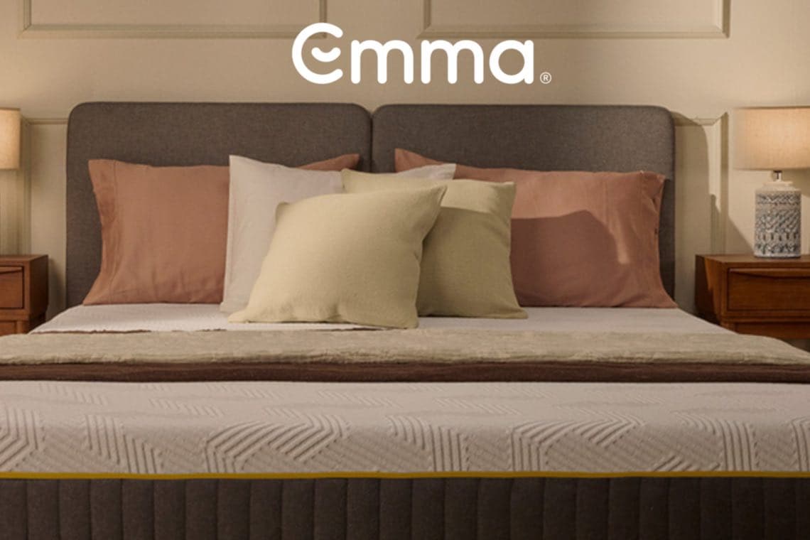 Emma Logo über Doppelbett mit Kopfteil, mehreren Kissen und Nachttischen mit Lampen links und rechts