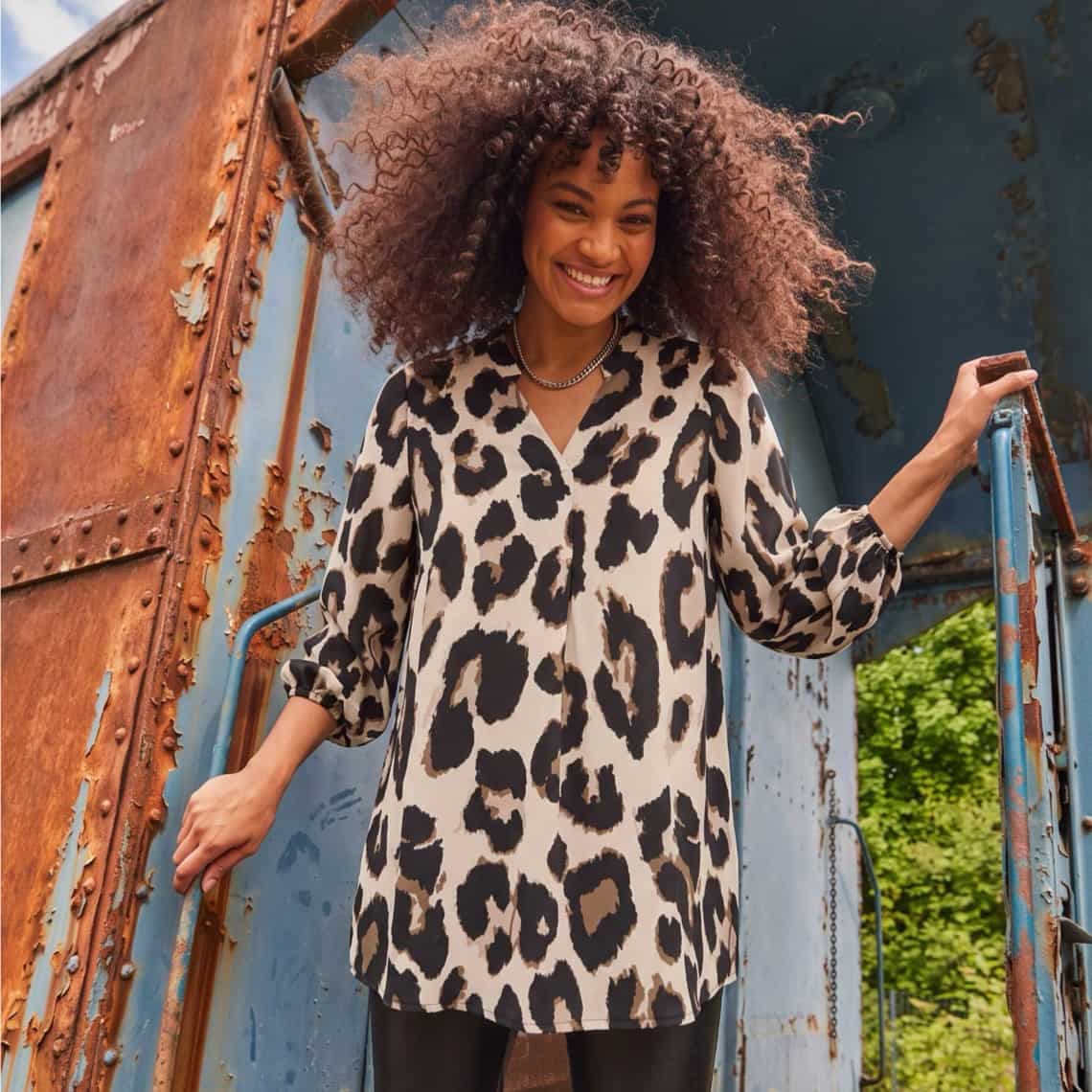 Frau in weiter Tunika mit großem Leo-Muster steht auf einer alten, rostigen Metalltreppe im Freien