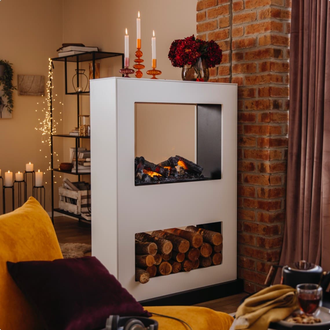 Weißer Kamin mit sichtbarem Feuer und Holzfach, davor gelbes Sofa, im Hintergrund Regal, Kerzen und Lichterkette