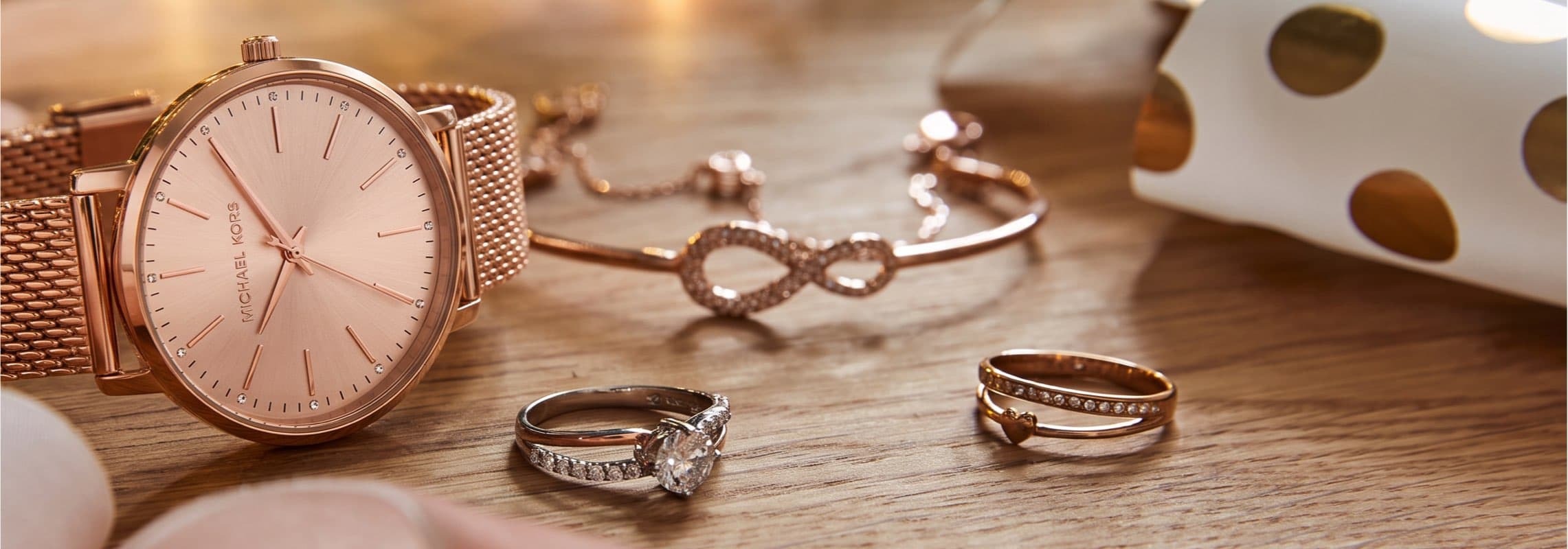 Roségoldene Armbanduhr neben Armband mit Unendlichkeitszeichen und zwei Ringen auf Holztisch, Geschenkpapier im Hintergrund