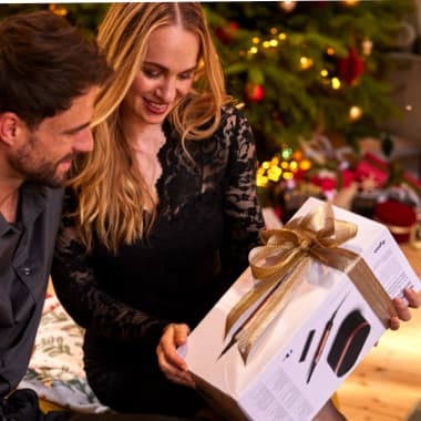 Paar sitzt vor Weihnachtsbaum, Frau hält großes Geschenk mit goldener Schleife und lächelt den Mann neben ihr an