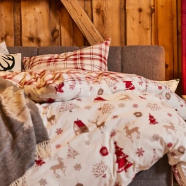 Bett mit Decke im Wintermotiv mit Hirschen und Tannen, dazu kariertes Kopfkissen vor rustikaler Holzwand