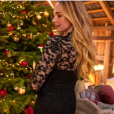 Frau mit langen, offenen Haaren im schwarzen Spitzenkleid steht seitlich vor geschmücktem Weihnachtsbaum im Wohnzimmer