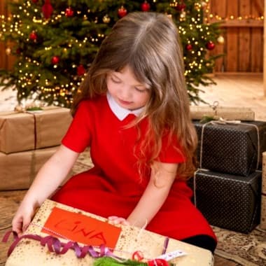 Mädchen im roten Kleid sitzt vor Weihnachtsbaum und hält ein Geschenk mit Namensschild „Lina“ in den Händen
