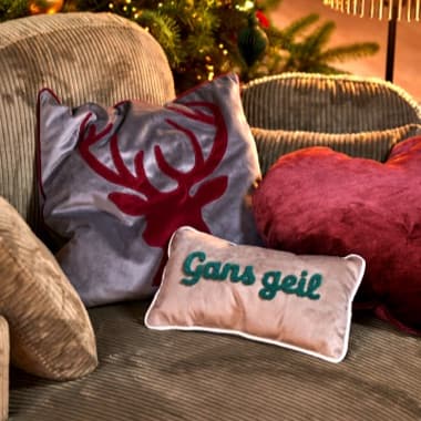 Kissen mit Aufschrift „Gans geil“ vor Kissen mit rotem Hirschkopf auf Sofa, dahinter Weihnachtsbaum