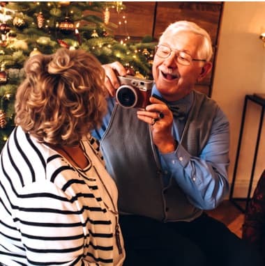 Älterer Mann fotografiert lachende Frau vor geschmücktem Weihnachtsbaum im Wohnzimmer