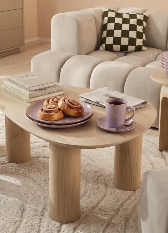Wohnzimmer mit ovalem Couchtisch, Zimtschnecken auf Teller und Tasse Kaffee vor Sofa mit kariertem Kissen