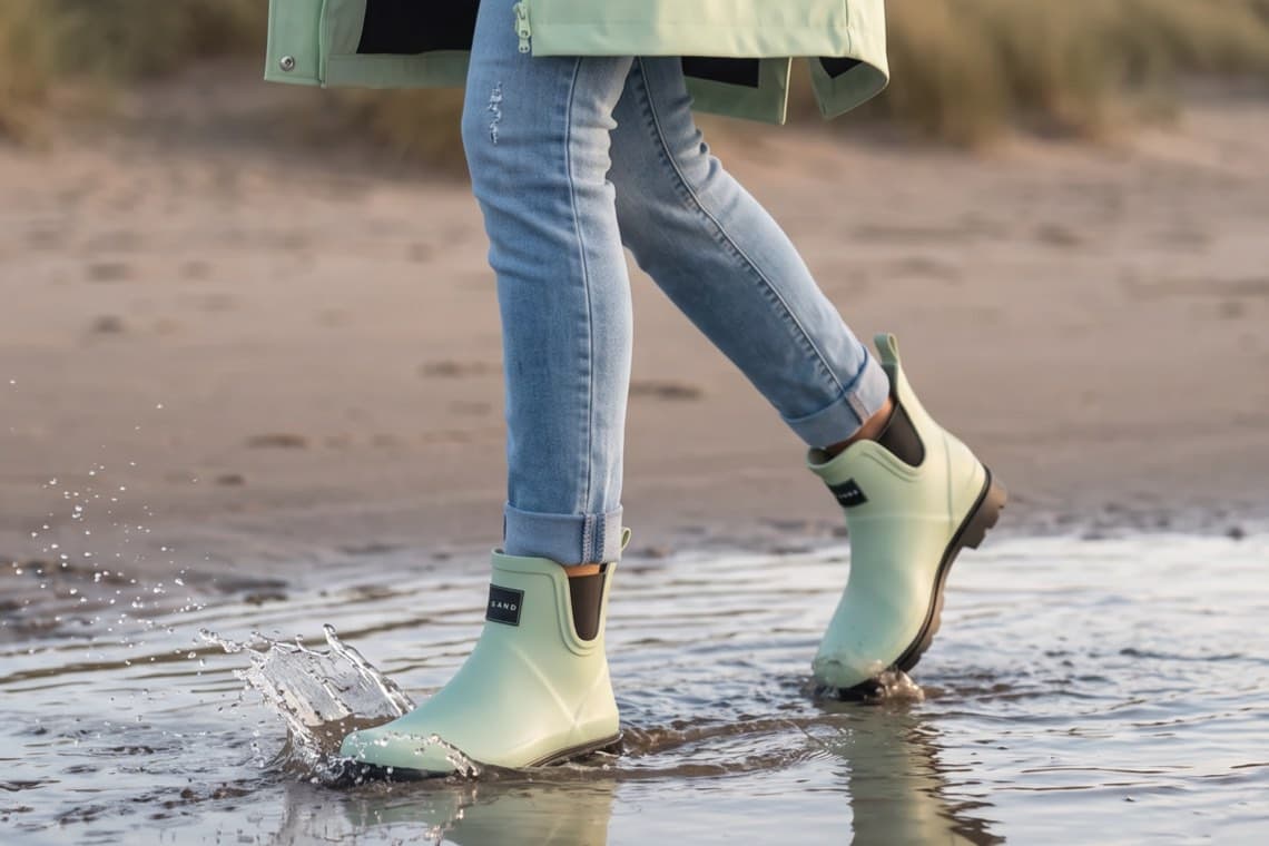 Person in hellgrünen Gummistiefeletten und Jeans läuft am Strand durch eine flache Wasserpfütze