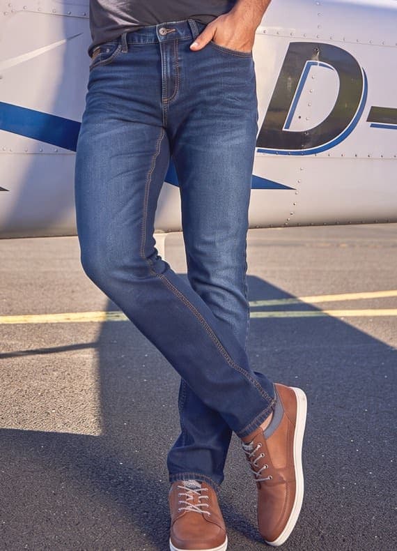 Mann in dunkelblauer Jeans und braunen Schnürschuhen lehnt lässig an einem Flugzeug auf dem Rollfeld