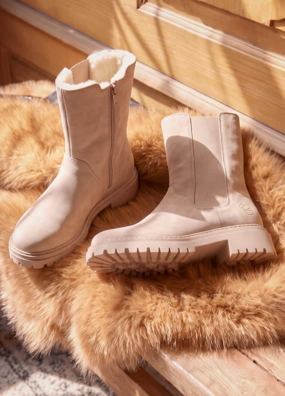 Beige Winterstiefel mit Profilsohle, ein Stiefel stehend, der andere liegend auf einem flauschigen Fell vor Holzoberfläche