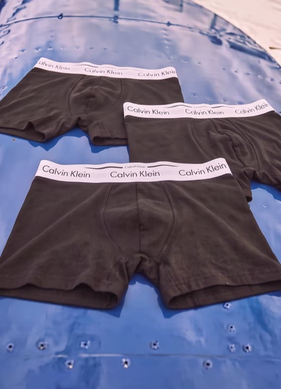 Drei schwarze Herren-Boxershorts mit weißem Calvin-Klein-Bund liegen nebeneinander auf glänzend blauem Untergrund