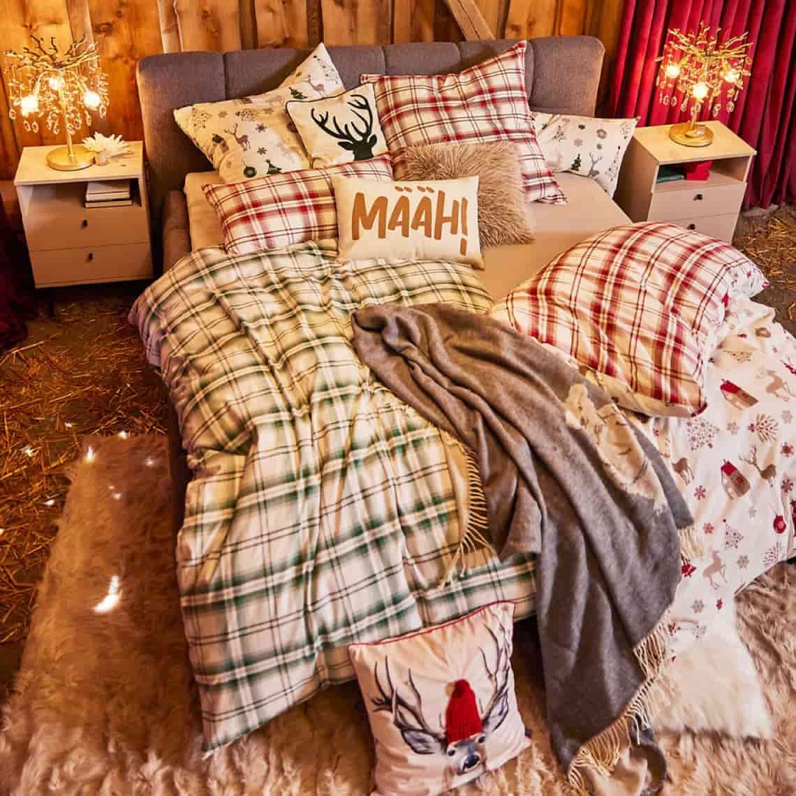 Kissen mit Aufdruck „MAÄH!“ auf winterlich dekoriertem Bett mit karierten Bezügen, Decke und Lichterbäumen am Rand