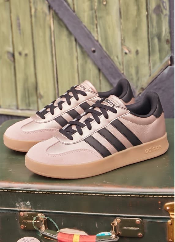 Bordeauxfarbene Sneaker mit hellen Streifen stehen seitlich auf einem alten Koffer vor Holzwand