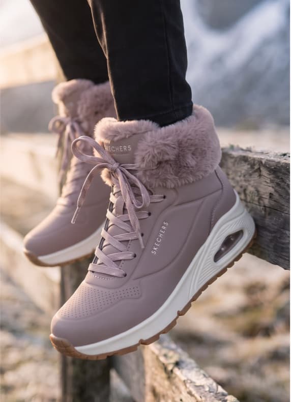 Rosa Skechers-Sneaker mit Fellrand an Füßen einer Person, die auf einem Holzzaun im Freien steht