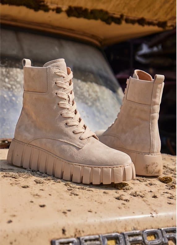 Beige Schnürstiefel mit dicker Profilsohle stehen auf einer staubigen Motorhaube vor einer schmutzigen Windschutzscheibe