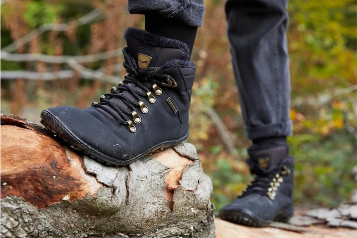Person mit schwarzen Wanderstiefeln steht auf einem Baumstamm im Wald, Fokus auf linkem Schuh und Schnürung