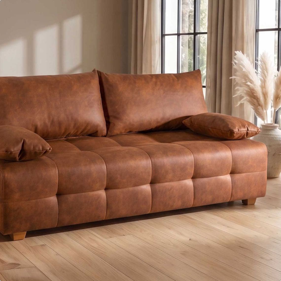 Braunes, gepolstertes Sofa mit zwei Kissen vor hohen Fenstern und hellen Vorhängen auf hellem Holzfußboden
