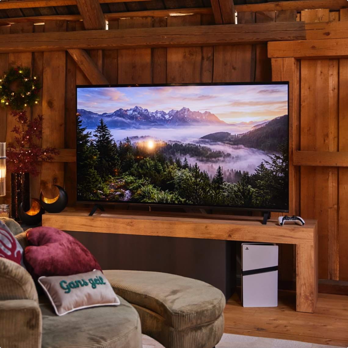 Großer Fernseher mit Berglandschaft steht auf Holzboard, davor Sofa mit Kissen, daneben Konsole und Controller