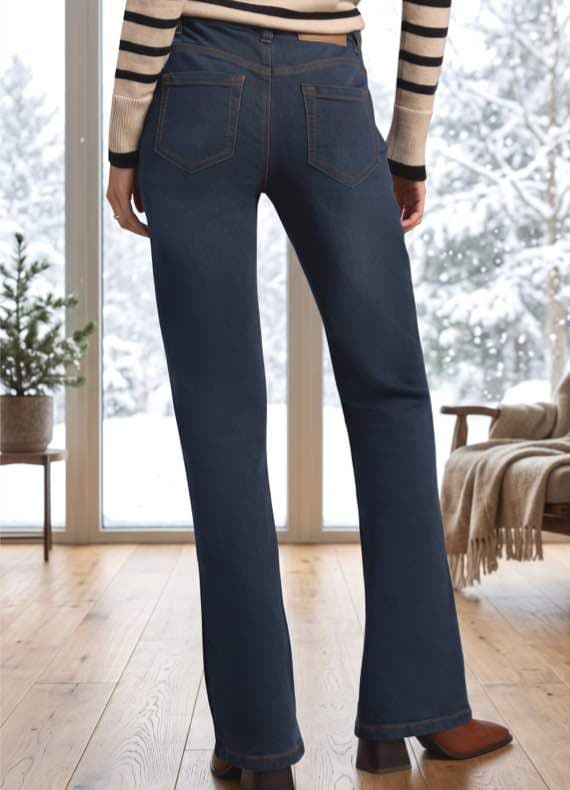 Frau von hinten in dunkelblauer Schlagjeans, gestreiftem Pullover, steht in hellem Wohnzimmer mit Blick auf verschneiten Garten