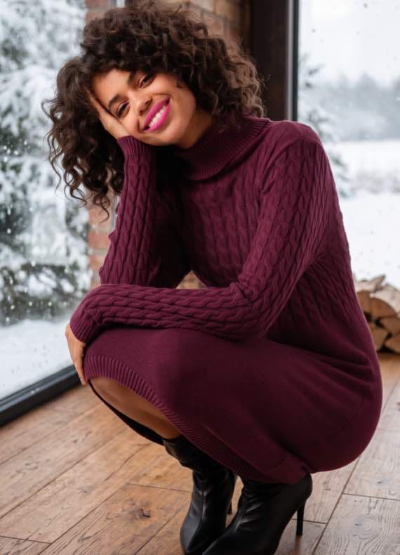 Frau in bordeauxrotem Strickkleid hockt am Fenster mit Winterlandschaft und legt den Kopf in die Hand