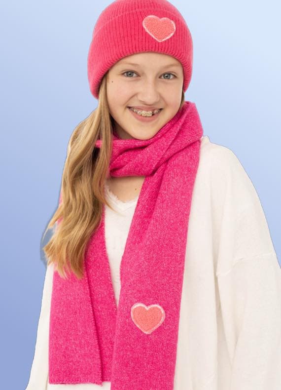 Mädchen mit Zahnspange trägt pinke Mütze und Schal mit Herzmotiven über weißem Oberteil vor blauem Hintergrund